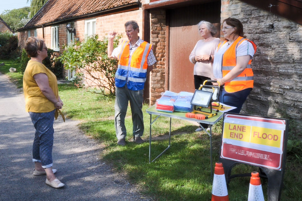 SRA community engagement support officer Dawn James talking to Derek Johnson, Jenny Pike and Sarah Elliott about the work of Ham Village Flood Defence Committee, with some of the committee's equipment laid out nearby.
