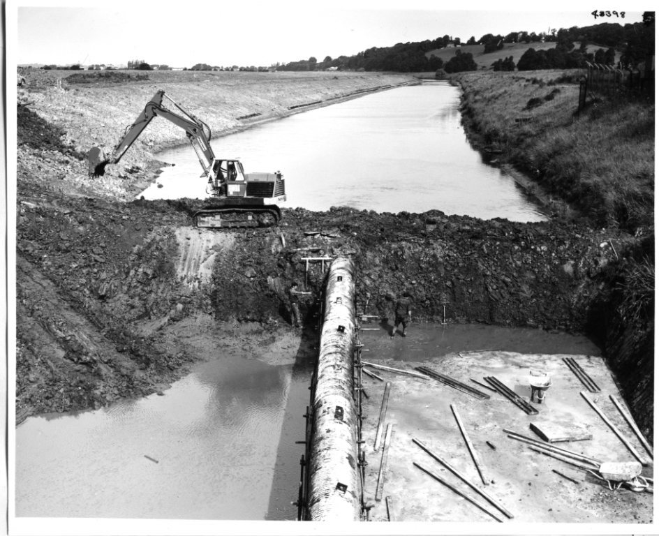 A black and white archive photo of an excavator working on top of a large earth dam across King's Sedgemoor Drain at Crandon, with lots less water on one side than the other.