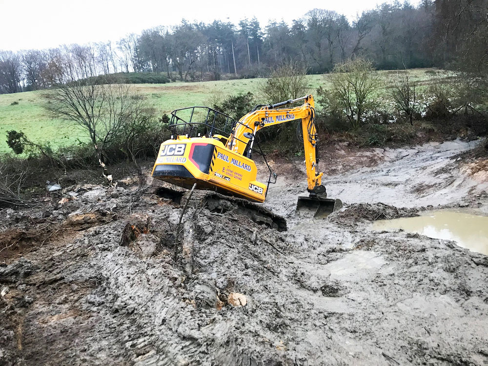 An excavator at an angle on a bank about to scoop silt from a pond restored at Witham Friary.