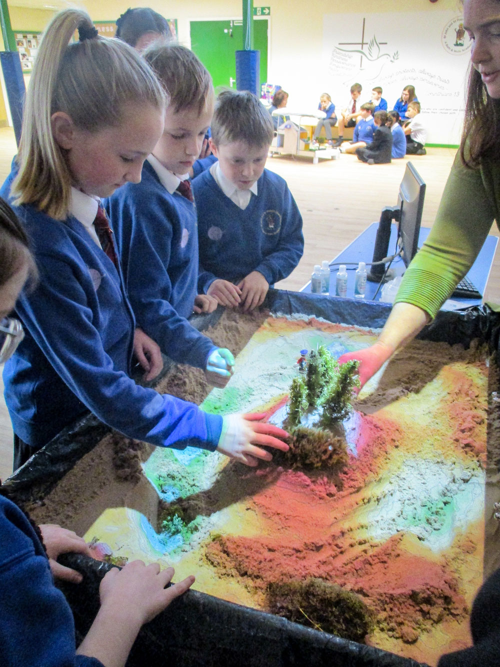 Pupils watch intently as they shape a tree-top mound in an augmented reality sandbox, with Emma Giffard discussing the effects created.
