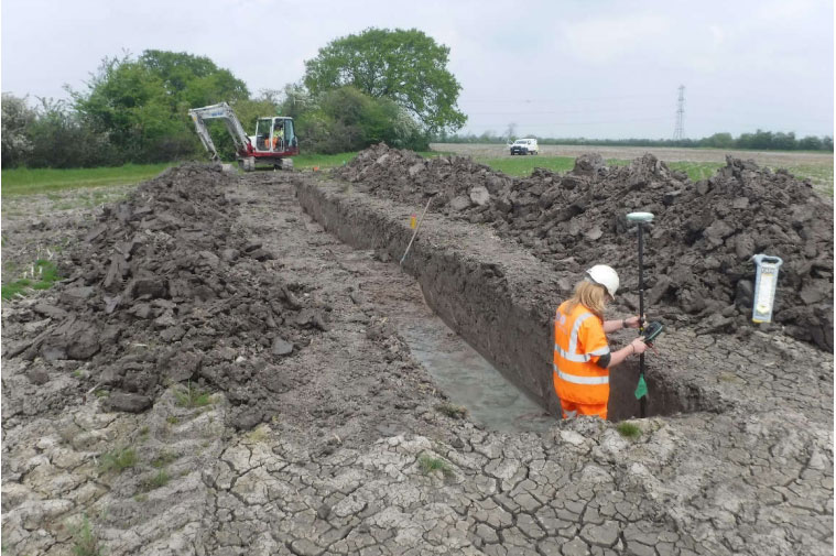 A blonde haired archaeologist using equipment in a trench with an excavator at the end of the trench.