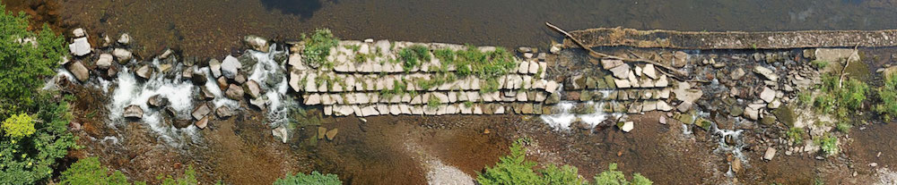Part of Dulverton Weir seen from above, with some loose stones and vegetation growing, and the river flowing over sections.