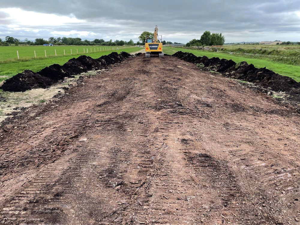 Compressed earth shaped into a bank, with topsoil stacked either side, and a tracked excavator at the far end of the bank.