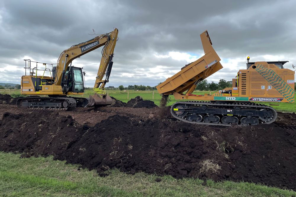 Two machines on top of a bank being built, one depositing material from its dumper, one using its arm and bucket to lay material.