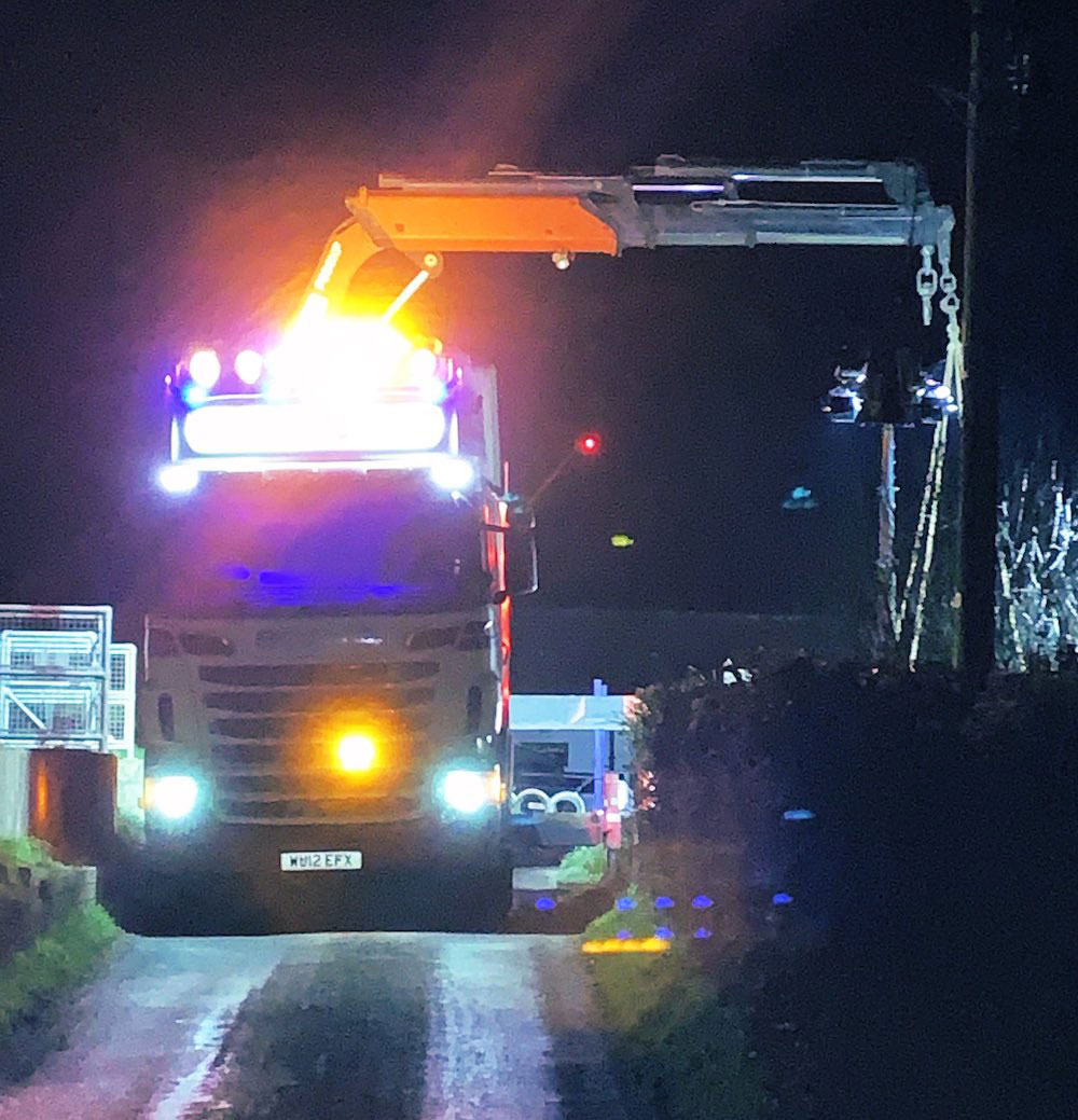 A brightly-lit lorry on road at night with crane on back lifting equipment at Elson's Clyse.
