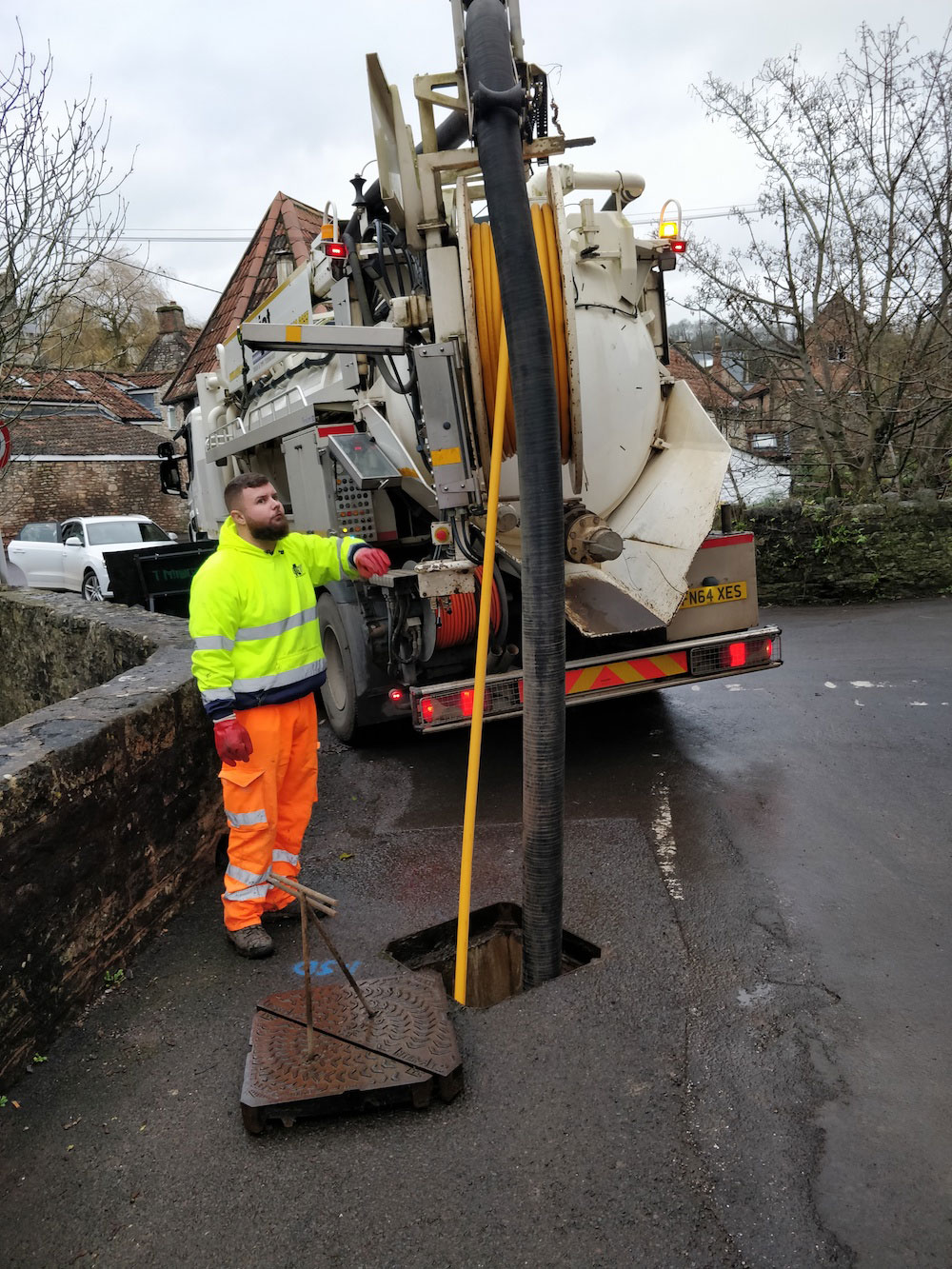 Worker in hi-vis clothing looking at a screen at the bak of a large drain jetting vehicle, from which two pipes are going down a drain.