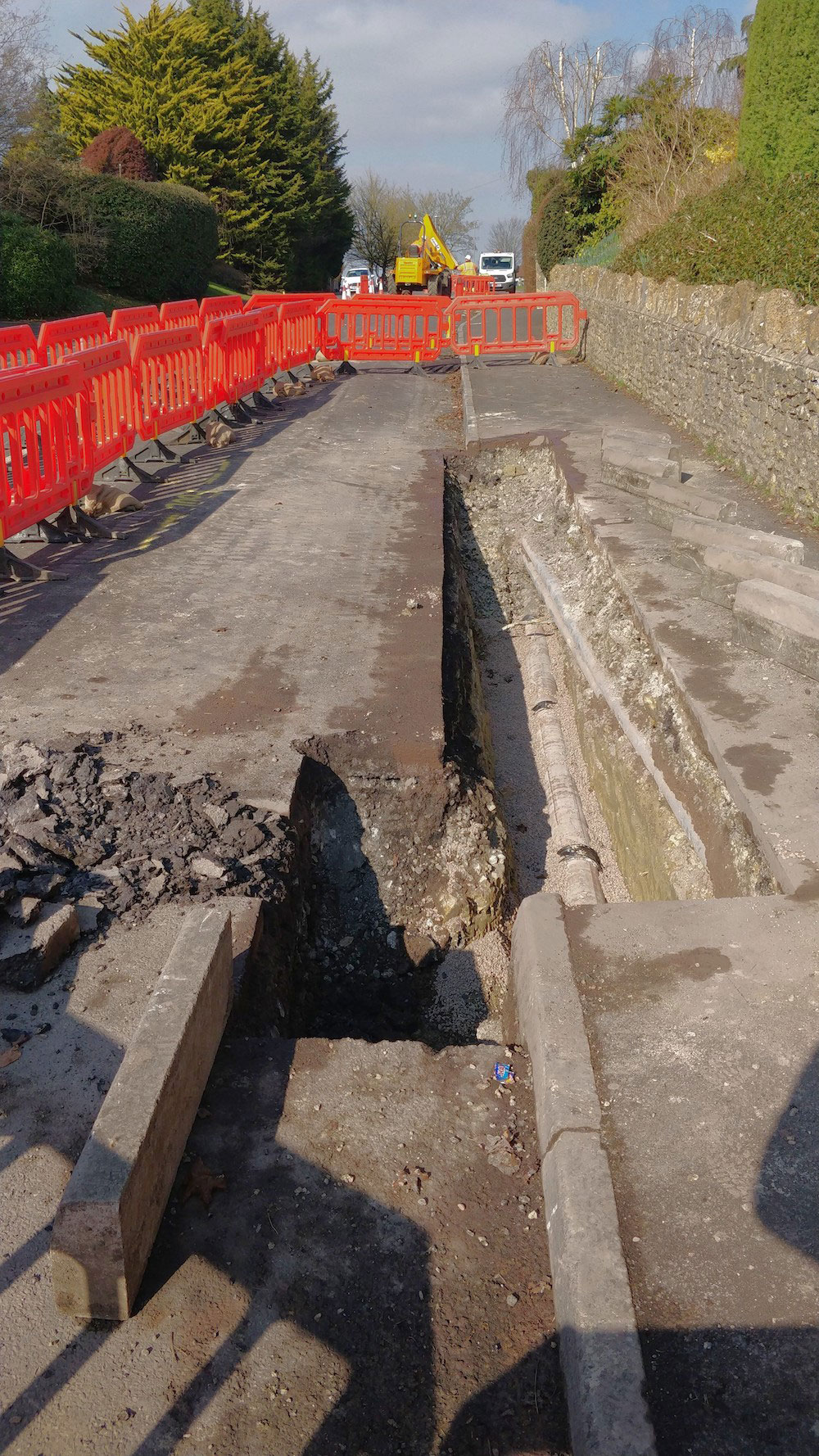 Sections of road and pavement dug up in neat trenches, exposing a pipe, with red barriers around the site.