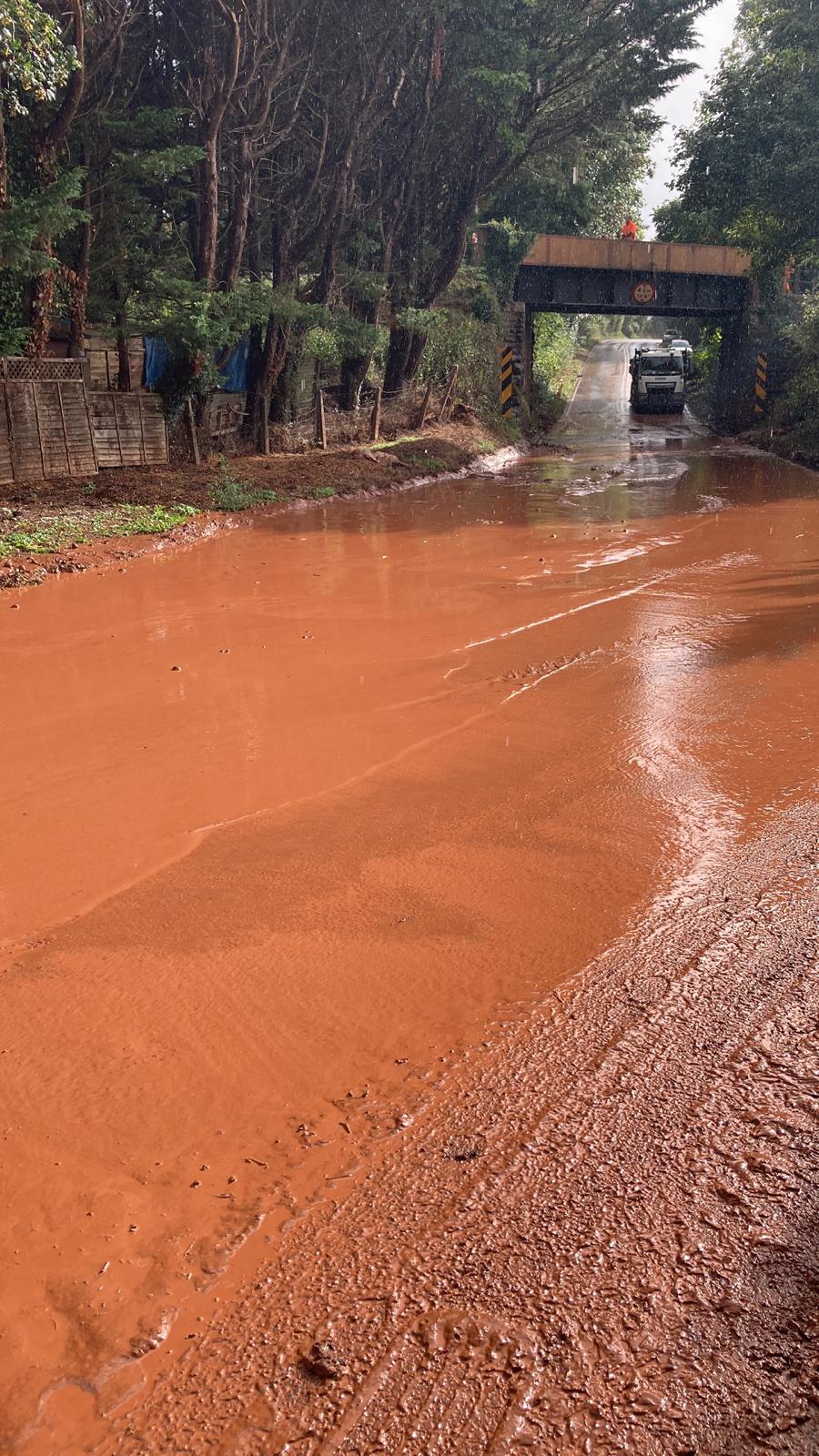 The A358 covered across both lanes with bright liquid mud, and a lorry stopped beyond a bridge.