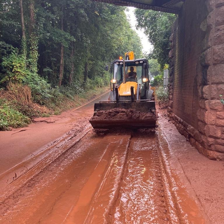 A digger in thick mud under a bridge on the A358 near Combe Florey, with its front bucket full of scooped-up mud.